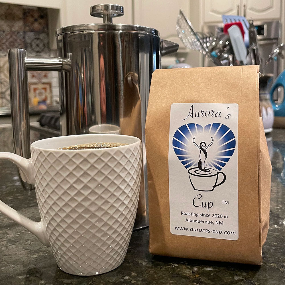 A stainless steel French press in the back ground, a bag of Aurora's Cup Coffee and a white ceramic mug.